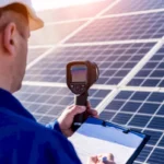 Eine Person mit Schutzhelm benutzt ein Handger&auml;t, um Solarmodule f&uuml;r ein PV-Gutachten zu pr&uuml;fen. Sie h&auml;lt ein Klemmbrett in der Hand und steht vor mehreren Reihen von Solarmodulen im Freien.