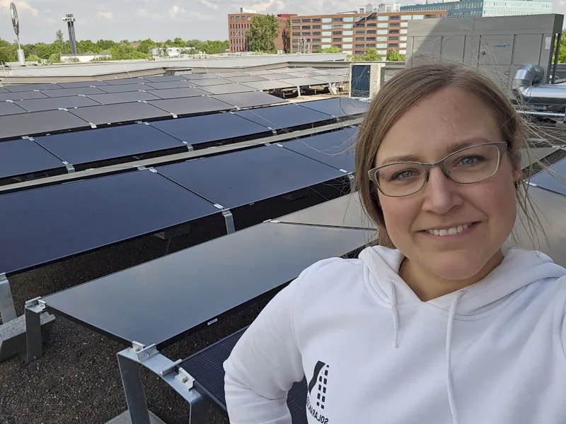 Eine Frau mit Brille und weißem Kapuzenpulli lächelt in die Kamera, während sie vor Reihen von PV-Gutachten - Solargutachten auf einem Dach steht. Im Hintergrund sind Gebäude und Grünflächen zu sehen.